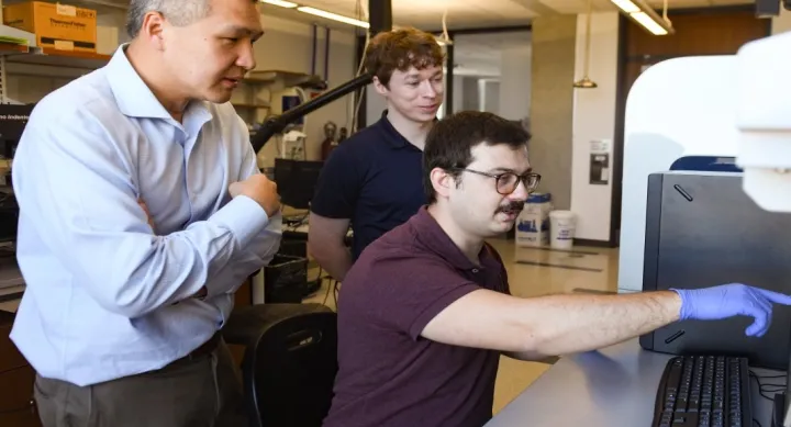 Professor Chih-Hao Chang, Mehmet Kepenekci, and Andrew Tunell wear business casual clothing and stand in front of a computer in the lab.