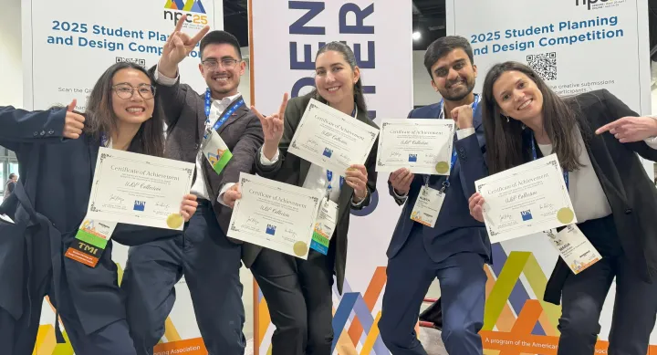 Five students in professional attire hold their certificates of achievement and make the hook-em horns sign in front of freestanding banners for the Student Planning and Design Competition.