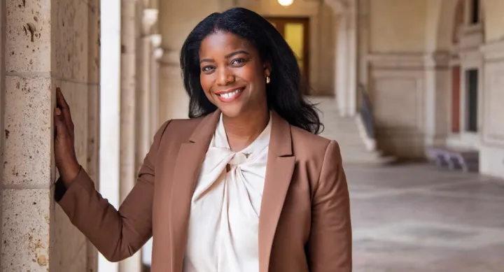 Nina Means wears a white blouse and a brown suit jacket and stands in the entryway to the UT Tower. She smiles and poses with her hand resting on one of the brick pillars.