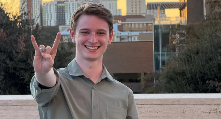Colby Jones wears a grey shirt and stands on a balcony overlooking a city, making a hook-em horns sign towards the camera.