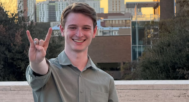 Colby Jones wears a grey shirt and stands on a balcony overlooking a city, making a hook-em horns sign towards the camera.