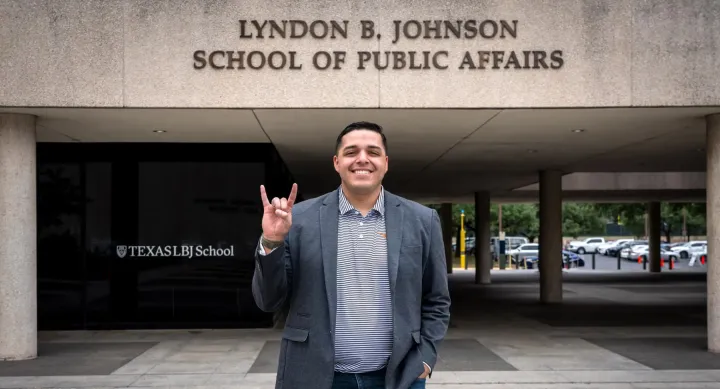 Steven Santoyo wears a grey suit jacket and makes the Hook 'em Horns hand sign in front of the Lyndon B. Johnson School of Public Affairs building.