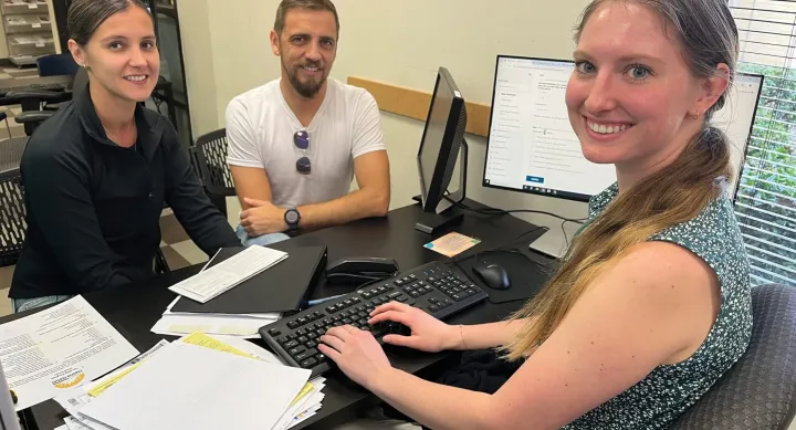 A woman sits at a desk full of tax forms and a keyboard. In front of her are a man and a woman. All three of them smile at the camera.