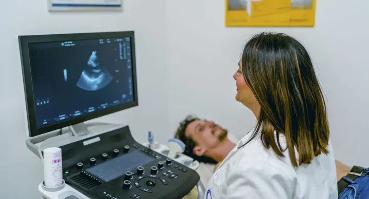 A woman in a lab coat examines an ultrasound image on a computer while a man lays behind her on a table.