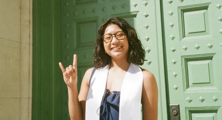 A woman in glasses stands in front of a bright green double door, making the hook 'em horns sign while wearing a white commencement sash.