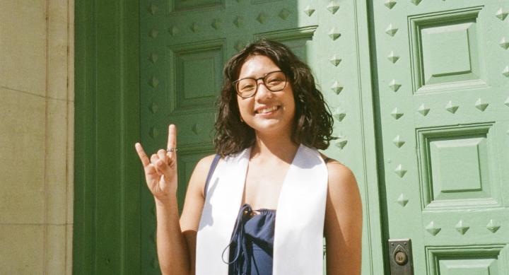 A woman in glasses stands in front of a bright green double door, making the hook 'em horns sign while wearing a white commencement sash.