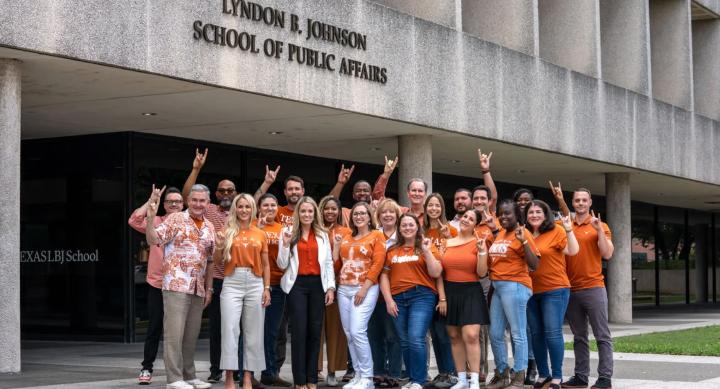 A group of students and faculty wear orange University of Texas at Austin shirts and make the Hook 'em horns sign in front of the Lyndon B. Johnson School of Public Affairs building.