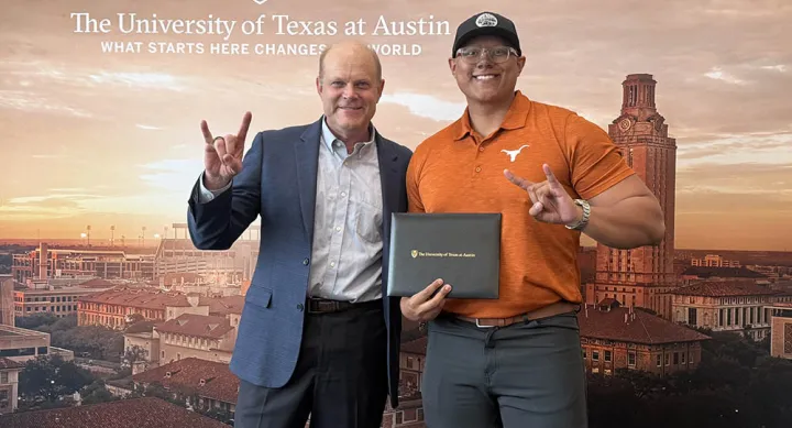 Dario Garcia wears an orange Longhorn polo and baseball cap, holding his diploma. He stands next to Eric Bickel, who wears a blue suit jacket. They make a Hook 'em horns sign with their hands in front of a University of Texas at Austin banner.