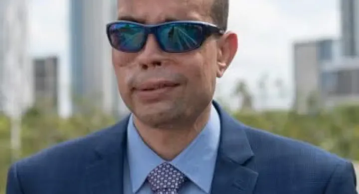 Qusay Hussein wears sunglasses and a blue suit. Behind him is a city skyline.