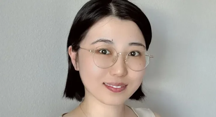 A woman wears glasses, a gold necklace, and a beige shirt and stands in front of a white wall.