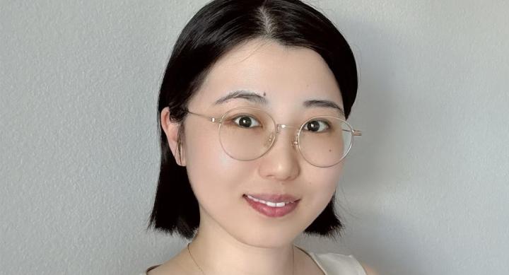 A woman wears glasses, a gold necklace, and a beige shirt and stands in front of a white wall.