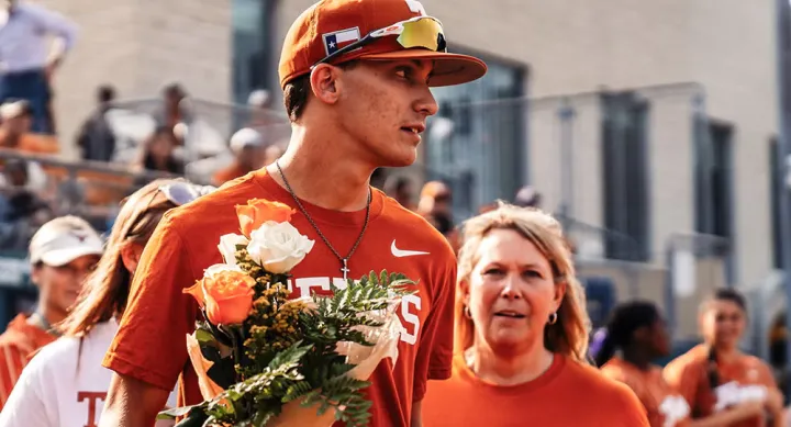 A man in an orange Texas shirt and orange Texas baseball cap holds a bouquet of orange and white flowers. Surrounding him are people in orange and white UT-Austin shirts sitting on bleachers.