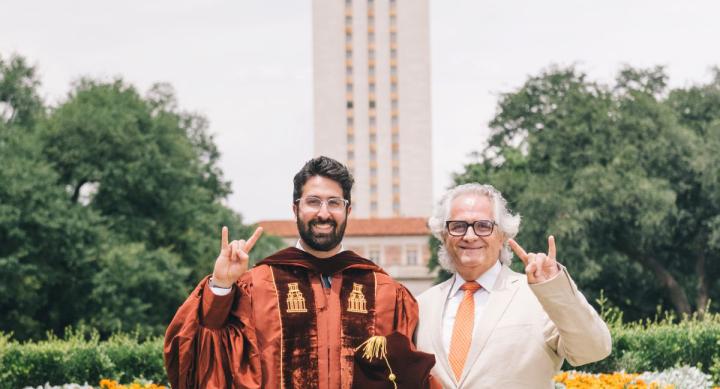 A man in a red graduation robe stands next to a man in a white suit and red tie. They make Hook 'em Horns hand signals in front of the UT Tower.