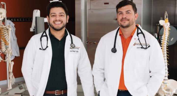 A man in a black shirt and a man in an orange shirt stand next to each other. Both wear a white doctor's coat and a stethoscope around their neck.