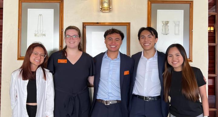 Five people wear businesswear and smile in front of framed artwork.