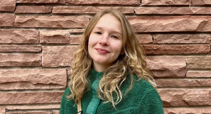 Ann Laudick A woman wears a green sweater and smiles in front of a red stone wall.
