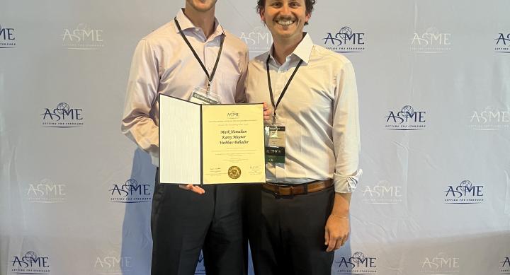 Two men in business casual hold a certificate and smile in front of an ASME banner.