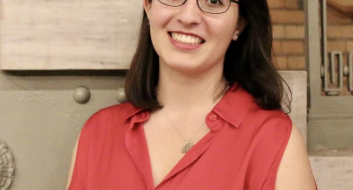 A woman wears glasses and a red blouse and stands in front of a staircase.