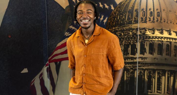A man in an orange shirt stands in front of a mural depicting a capitol building and an American flag.