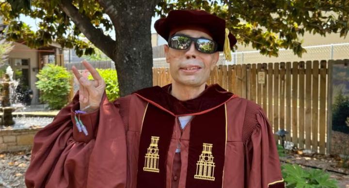 A man wears a red doctoral convocation gown and makes the Texas Hook'em hand gesture in an outdoor setting.