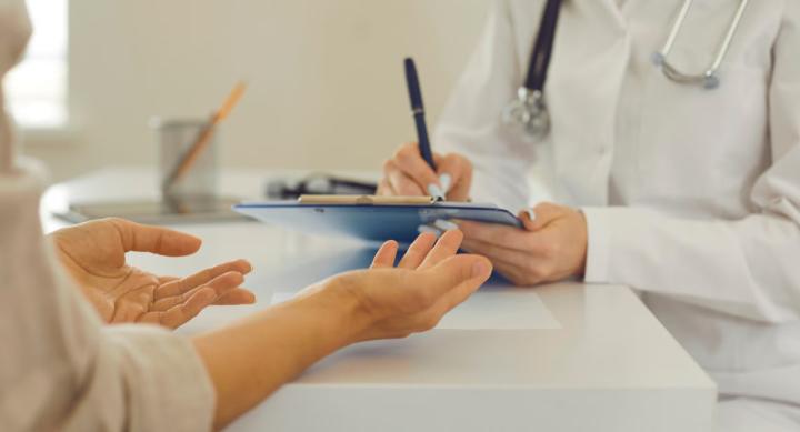 A doctor in a lab coat writes notes on a tablet while speaking with a patient at a white table.