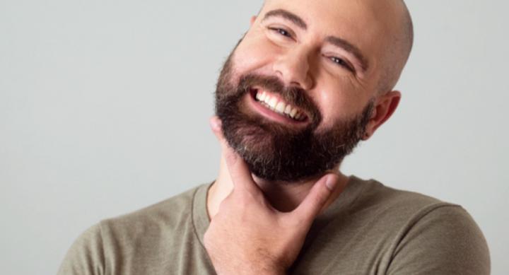 A man smiles and wears a brown t-shirt in front of a grey background.