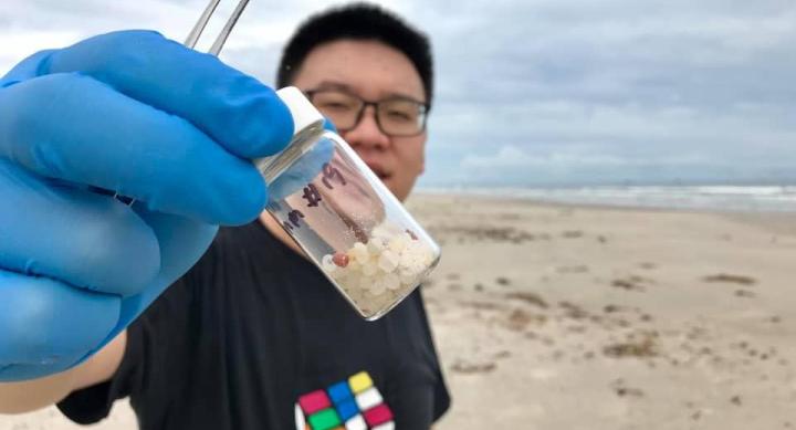 A man stands on a beach holding a glass vial of white orbs in his hand, which wears a blue medical glove.