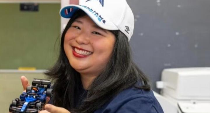 A woman wears a baseball cap and holds up a toy car in an indoor space.