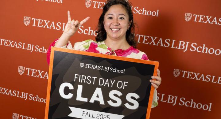 In front of an orange LBJ School photo banner, a woman makes the Hook 'em sign holding a sign that says "First Day of Class Fall 2025."