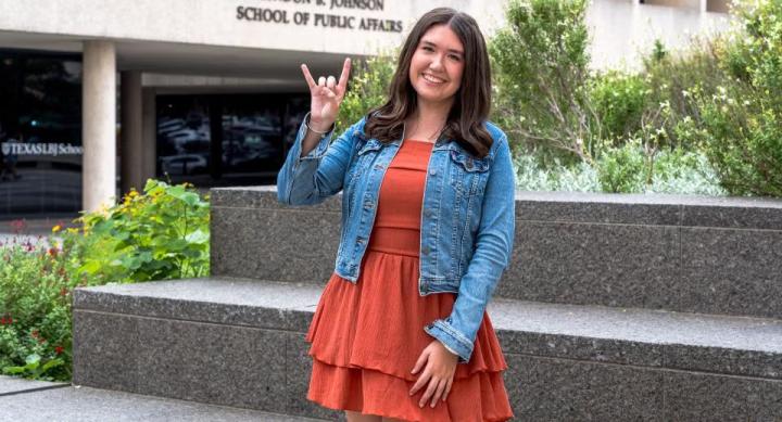 A person wears an orange dress and a jean jacket and makes a "Hook 'em Horns" hand sign in front of an academic building.