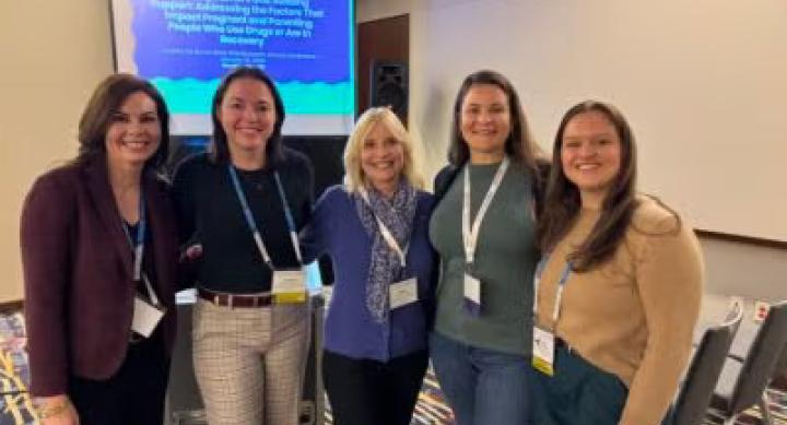 Five people in business casual wear lanyards and stand in front of a powerpoint display.