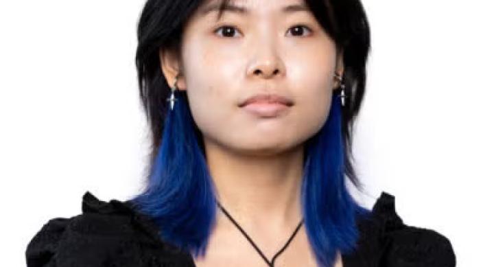 A person wears a black blouse and stands in front of a white background.