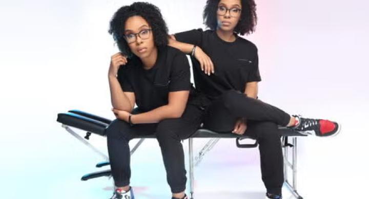 Twins in black clothes sit next to each other on a massage table against a white background.