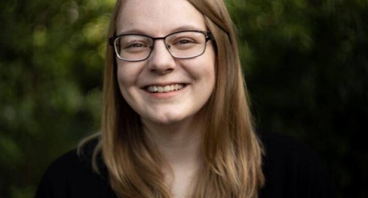 A person wears glasses and a black shirt and smiles in front of a forest background.