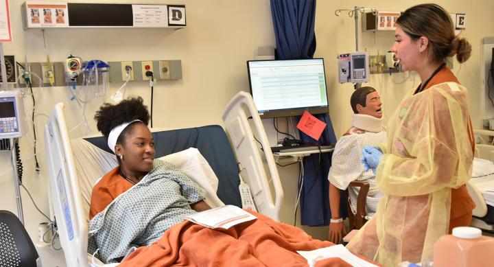 Two nursing students simulate a patient-nurse interaction in a hospital bed.