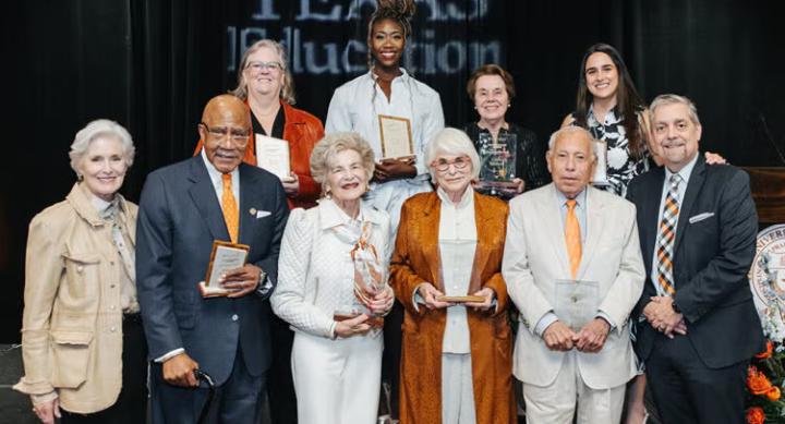 A group of people in formal wear pose with their award plaques on a stage.