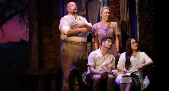 Four actors pose in 1940s era costumes onstage with the set of a brick building behind them.