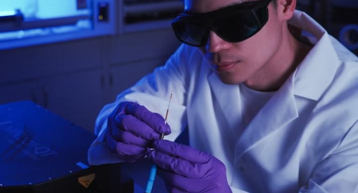 A researcher in a white lab coat, gloves, and goggles holds a probe.