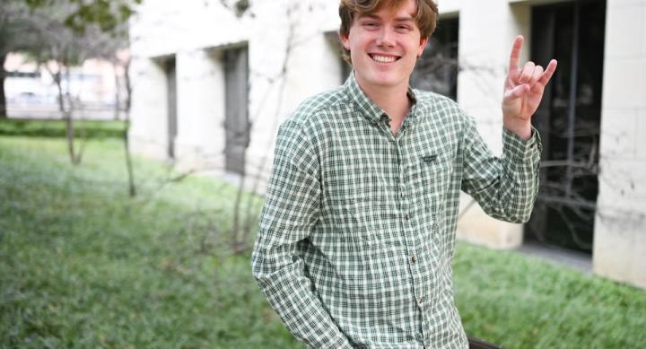 A person in a green shirt makes a Hook 'em hand sign in front of a campus building.