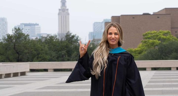 A person in graduation regalia makes a Hook 'em hand sign in front of the UT tower.