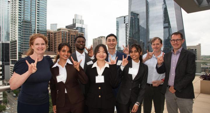 A group of students and professors in businesswear make the Hook 'em hand sign in an outdoor urban setting.