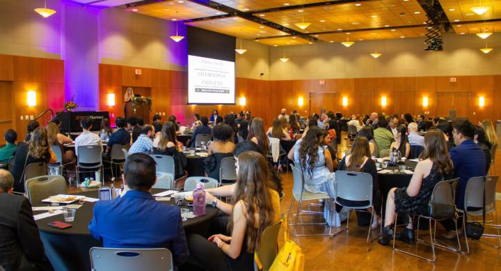 An audience sits at round tables with snacks and drinks in a conference room.