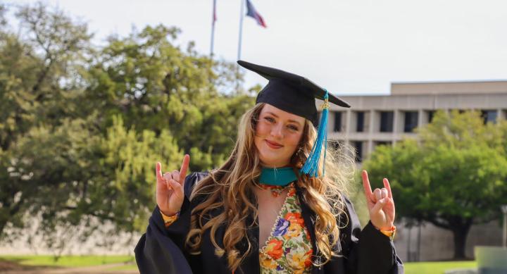 A person in graduation regalia poses in front of the LBJ library.