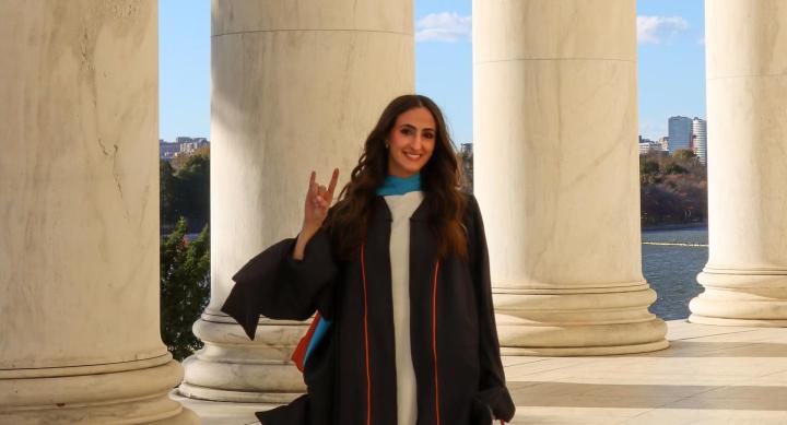 A person in graduation regalia makes a Hook 'em hand sign in front of white columns.