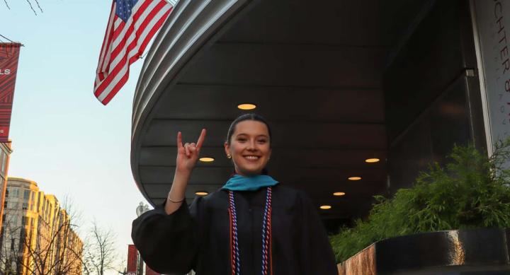 A person in graduation regalia stands in front of a building.