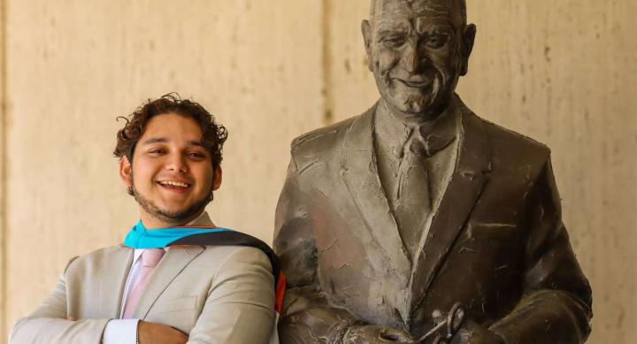 A man in a suit and a graduation hood stands next to a statue.