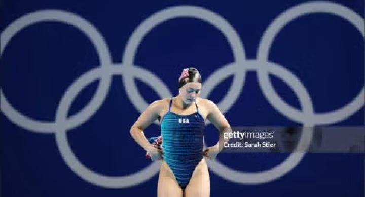 A person in a swimsuit and swimcap stands in front of a blue Olympic logo.