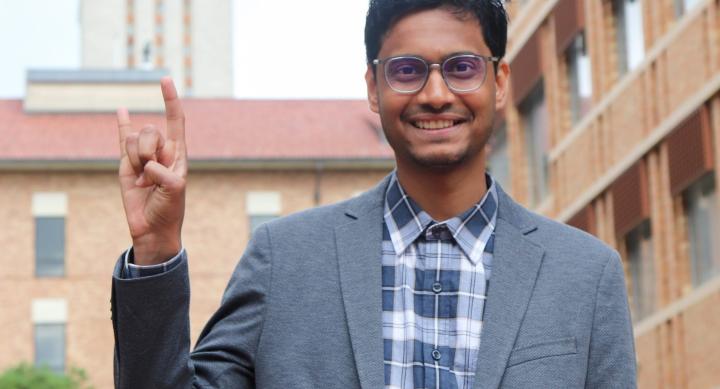 A person in a suit stands on campus in front of the UT Tower and makes a Hook 'em hand sign.
