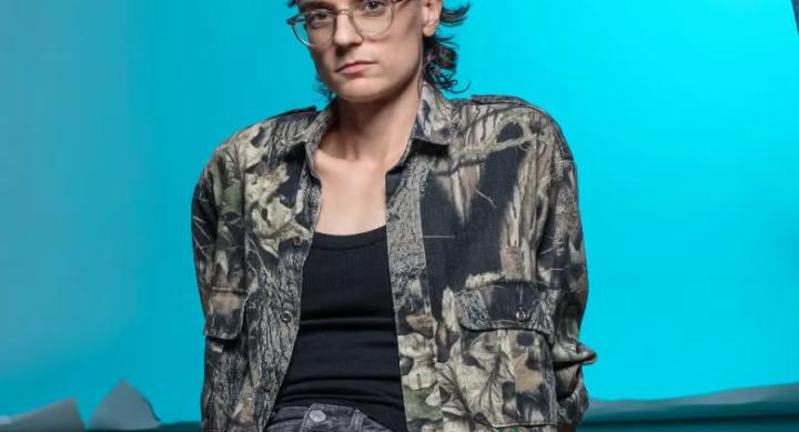 A person in a camo shirt sits on a floor in front of a blue background.