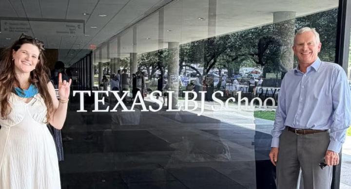Emma and Jim Niewald pose in front of a glass window depicting the Texas LBJ School logo.